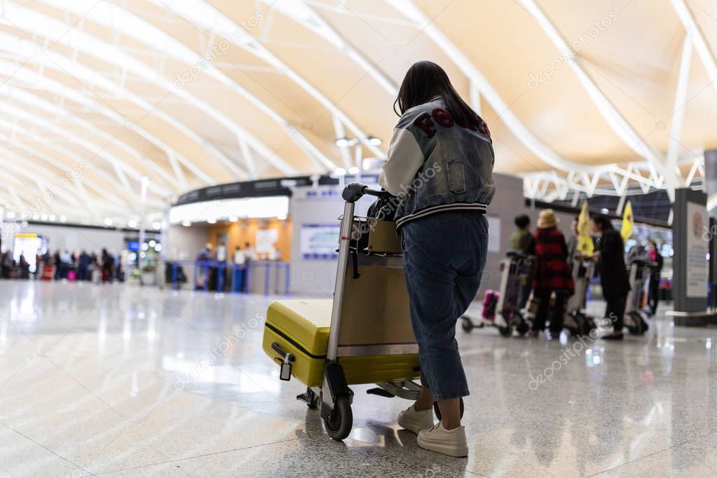 Young woman from behind transporting luggage from arrival parking to international airport departure terminal by luggage trolley