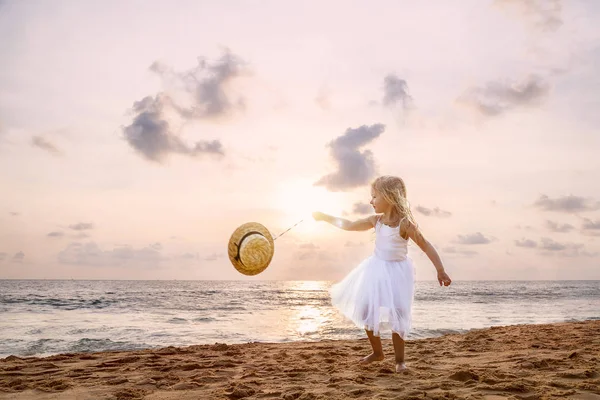 Menina bonito criança com cabelo loiro em um vestido branco tutu e chapéu andando em uma praia de areia ao pôr do sol. Memórias de infância feliz, férias de verão, conceito de viagem — Fotografia de Stock