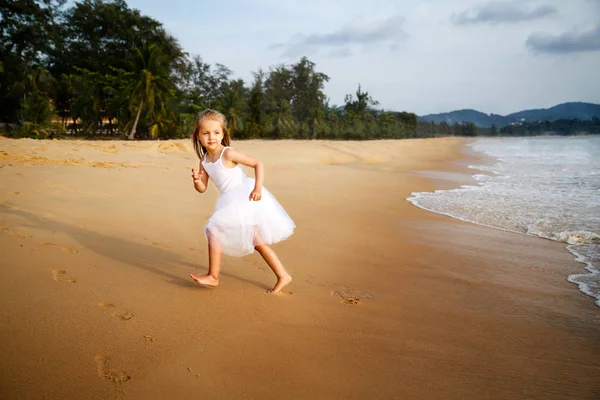 Menina bonito criança com cabelo loiro em um vestido branco tutu correndo em uma praia de areia ao pôr do sol. Memórias de infância feliz, férias de verão, conceito de viagem — Fotografia de Stock