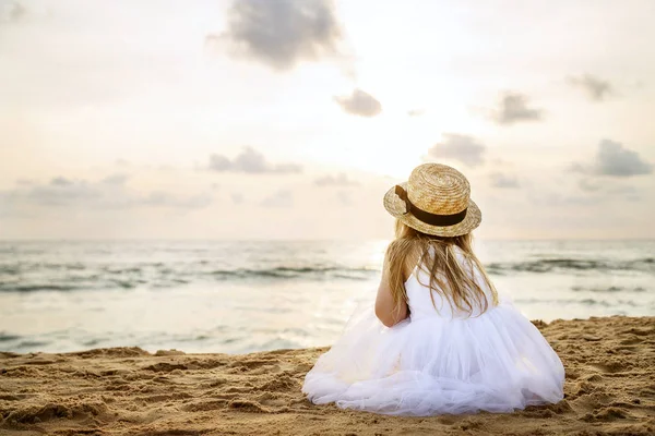 Menina bonita por trás com cabelo loiro longo em um chapéu de palha e um vestido de tutu branco sentado em uma praia de verão. Dia ensolarado, pôr do sol, céu dramático — Fotografia de Stock