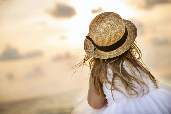 Menina bonita por trás com cabelo loiro longo em um chapéu de palha e um vestido de tutu branco sentado em uma praia de verão. Dia ensolarado, pôr do sol, céu dramático — Fotografia de Stock