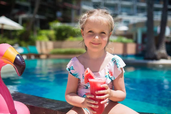 Menina bonito em maiô sentado perto da piscina com flamingo rosa inflável e beber suco de melancia fresca. Verão férias, relaxamento, conceito de viagem — Fotografia de Stock