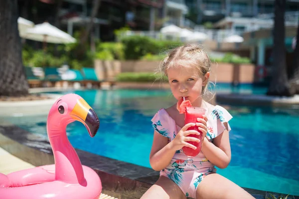 Menina bonito em maiô sentado perto da piscina com flamingo rosa inflável e beber suco de melancia fresca. Verão férias, relaxamento, conceito de viagem — Fotografia de Stock