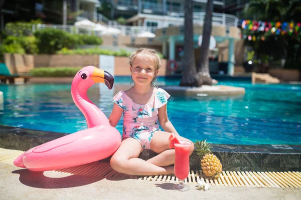Menina bonito em maiô sentado perto da piscina com flamingo rosa inflável, bola, suco de melancia fresca e abacaxi. Verão férias, relaxamento, conceito de viagem — Fotografia de Stock