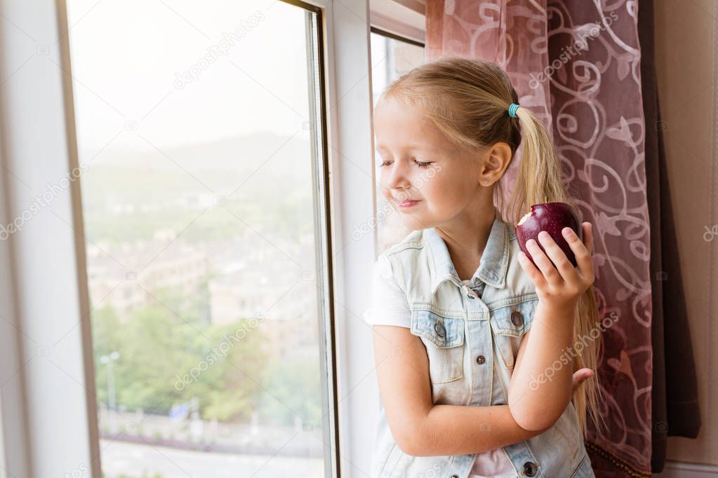 Pretty blonde little girl sitting on the sill at home, looking in window and holding red apple. Healthy food concept