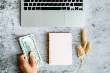 Masaüstü flatlay: laptop, Notebook, kadın elleri tutan 100 dolar faturaları gri mermer arka planda yatıyor. İş, finans konsepti