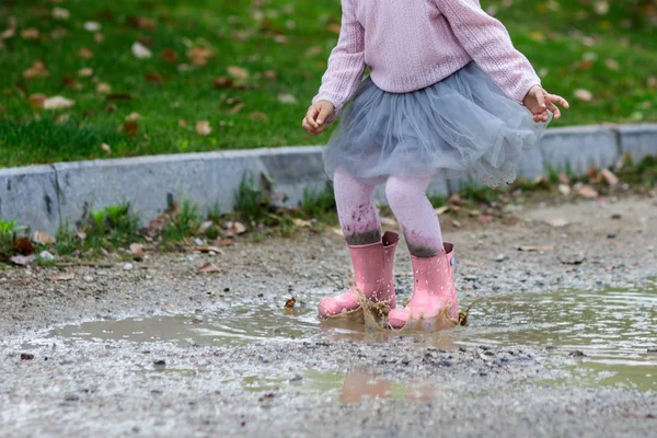 Little girl in rubber boots and tutu dress jumping in puddle. Water is splashing from kid feet as she is jumping and playing in the rain. — Stock Photo, Image