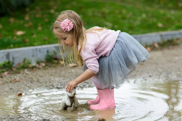 Beautiful little girl in a tutu skirt batting a teddy bear in a puddle on the street — Stock Photo, Image