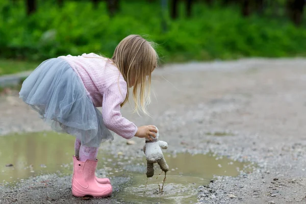 Beautiful little girl in a tutu skirt batting a teddy bear in a puddle on the street — Stock Photo, Image