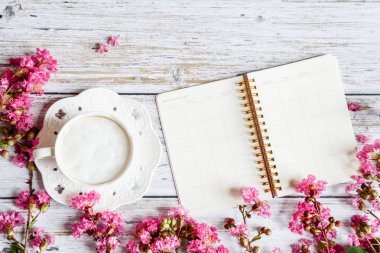 Kahvaltı için sabah kahve bardağı, defter, kalem, beyaz ahşap masa üzerinde pembe çiçekler, üst görünüm, düz yatıyordu tarzı. Kadın çalışma masası. Mockup, genel merkez