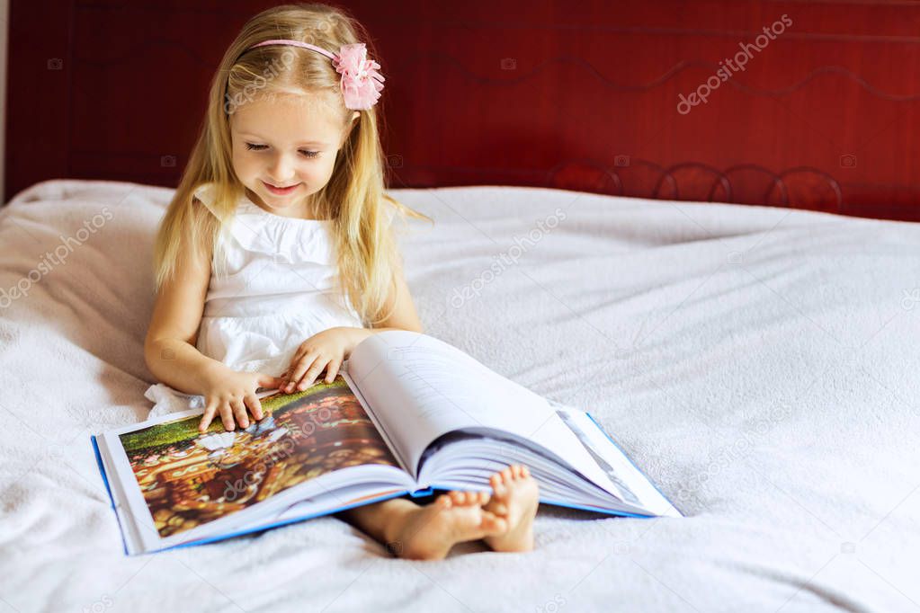 little girl with blonde hair reading book on the bed at home