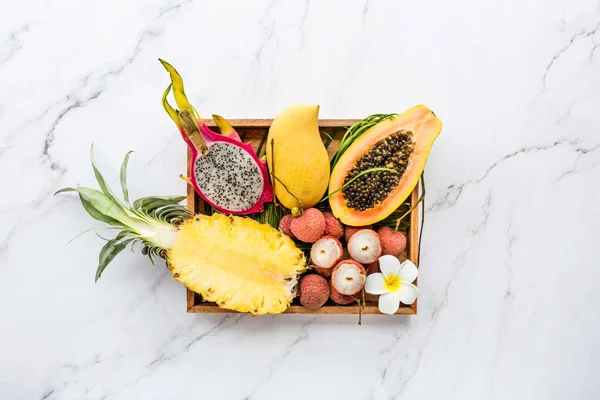 Fresh exotic fruits in wooden box on white marble background - sliced papaya, mango, pineapple, dragon fruit, lychee. Mockup, flat lay, overhead. Top view.