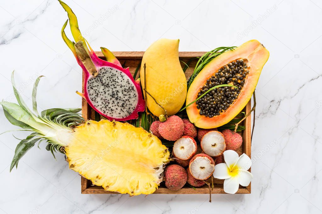Fresh exotic fruits in wooden box on white marble background - sliced papaya, mango, pineapple, dragon fruit, lychee. Mockup, flat lay, overhead. Top view.