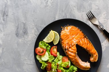 Kızarmış somon balığı bifteği, sebze salatası ve siyah tabakta limon. Balıktan, maruldan, domatesten, salatalıktan, salatalıktan, üst manzaradan, düz yataktan, maketten, genel masraftan oluşan yaratıcı bir düzen. Sağlıklı gıda konsepti