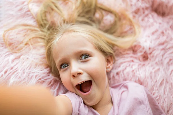 Милая маленькая девочка с длинными светлыми волосами, лежащая на розовом меховом одеяле и делающая селфи. Положительные эмоции, выражение лица, веселье. Счастливый улыбчивый малыш, день детства, образование, дети с использованием концепции технологии — стоковое фото