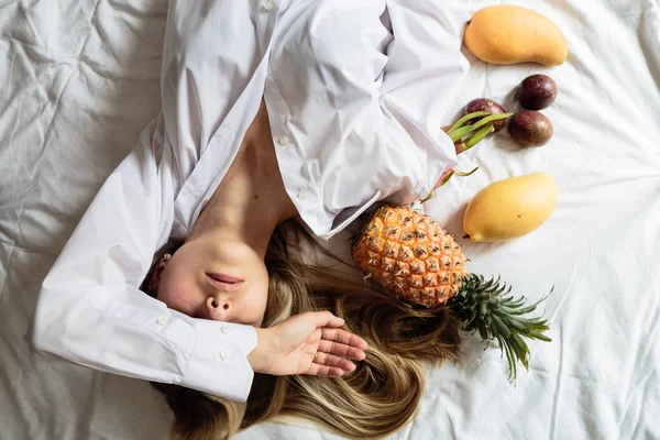 Junge Frau mit langen blonden Haaren in weißem Hemd auf dem Bett liegend. Sommer gesunde rohe exotische Früchte im Bett. Ansicht von oben, über Kopf. Zeit zum Entspannen und Träumen. Konzept der gesunden Ernährung — Stockfoto