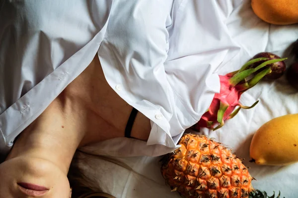 Junge Frau mit langen blonden Haaren in weißem Hemd auf dem Bett liegend. Sommer gesunde rohe exotische Früchte im Bett. Ansicht von oben, über Kopf. Zeit zum Entspannen und Träumen. Konzept der gesunden Ernährung — Stockfoto