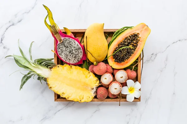 Fresh exotic fruits in wooden box on white marble background - sliced papaya, mango, pineapple, dragon fruit, lychee. Mockup, flat lay, overhead. Top view.