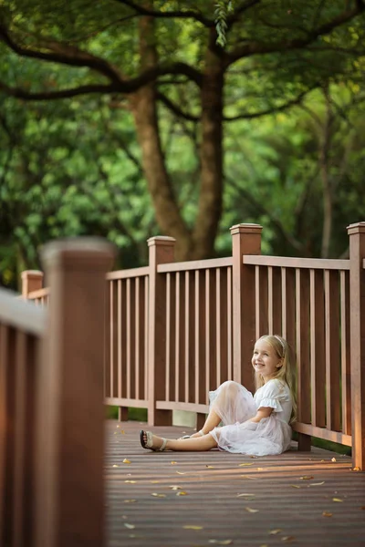 Adorable petite fille aux longs cheveux blonds assise sur un pont en bois dans un parc ensoleillé journée d'été. Magnifique enfant. Enfant souriant heureux, journée de l'enfance, concept d'éducation — Photo