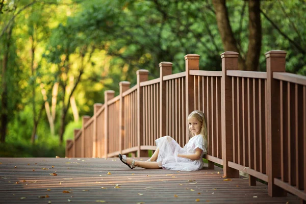 Αξιολάτρευτο κοριτσάκι με μακριά ξανθά μαλλιά κάθεται σε ξύλινη γέφυρα στο πάρκο ηλιόλουστη μέρα του καλοκαιριού. Όμορφο παιδί. Χαρούμενο χαμογελαστό παιδί, παιδική ηλικία, έννοια της εκπαίδευσης — Φωτογραφία Αρχείου
