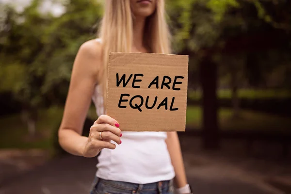 Conceito de igualdade de gênero como mãos de mulher segurando uma folha de papel com inscrição Somos iguais sobre um fundo de rua da cidade. Mulher protestando ao ar livre. Metáfora da questão social . — Fotografia de Stock