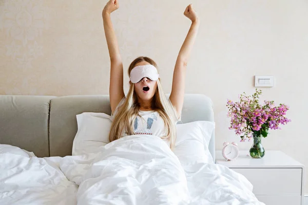 Glückliche junge blonde Frau in Pyjama und Augenbinde, die aufwacht, gähnt und sich im Bett dehnt und einen guten Tag hat. Morgenlicht im Schlafzimmer. Entspannung und gesundes Konzept — Stockfoto