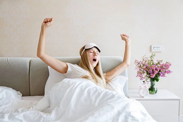 Glückliche junge blonde Frau in Pyjama und Augenbinde, die aufwacht, gähnt und sich im Bett dehnt und einen guten Tag hat. Morgenlicht im Schlafzimmer. Entspannung und gesundes Konzept — Stockfoto