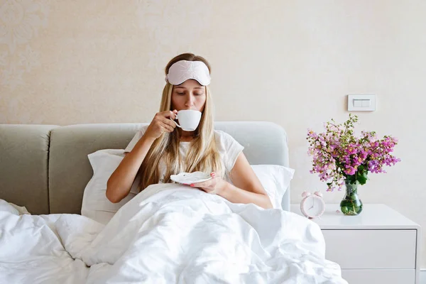 Schöne, glückliche junge Frau, die morgens nach dem Aufwachen im Bett Kaffee oder Tee trinkt. Porträt eines lächelnden blonden Mädchens in stylischem Pyjama, das ihren Drink genießt, während es sich zu Hause entspannt. — Stockfoto