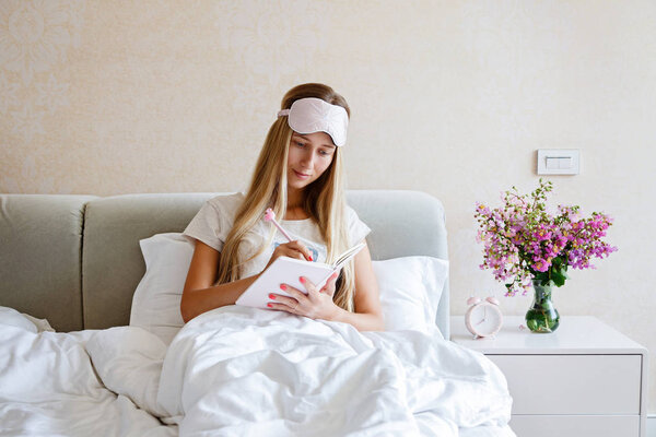 Beautiful Happy Young Woman writing something in notebook While Lying In Bed After Waking Up In Morning. Portrait Of Smiling Blonde Girl in stylish pajamas in bedroom