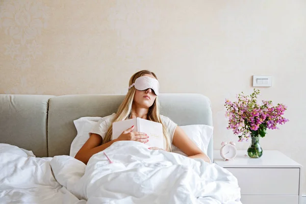 Schöne glückliche junge Frau, die mit Notizbuch im Bett schläft. Porträt eines stilvollen blonden Mädchens mit Augenbinde im Schlafzimmer. — Stockfoto