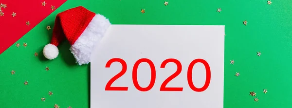 赤とコンフェッティで緑の背景にサンタの帽子と紙のブランク。新年とクリスマスのコンセプトウェブデザインのための水平バナー。グリーティングカード、クリスマスお祝い2020 。フラットレイモックアップ — ストック写真