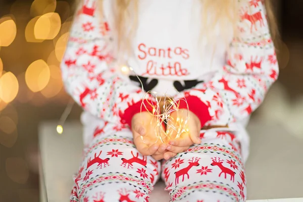 Petite fille mignonne avec de longs cheveux blonds en pyjama de Noël tenant guirlande à la maison. Décorations de Noël autour. Joyeux moments d'enfance. Concept vacances d'hiver — Photo