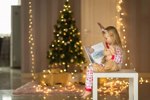 Petite fille mignonne avec de longs cheveux blonds en pyjama de Noël livre de lecture avec ours en peluche. Décorations de Noël autour. Joyeux moments d'enfance. Concept vacances d'hiver — Photo