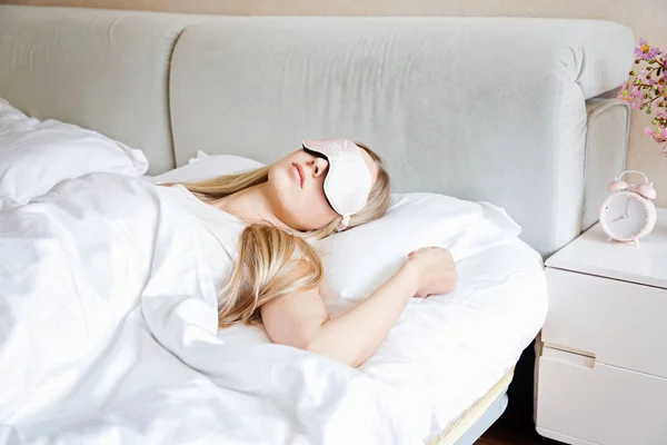 Young woman with long blonde hair sleeps on comfortable bed in a mask for sleeping. Blindfold on eye. Morning at home. White pillow and Blanket. Bedroom vibes