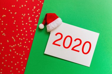 İçinde Noel Baba şapkası olan boş bir kağıt konfetiyle kırmızı ve yeşil arka plan. Noel ve yeni yıl konsepti. Kutlama kartı, xmas kutlaması 2020. Düz konum, şablon, üst görünüm, kopyalama alanı, modelleme