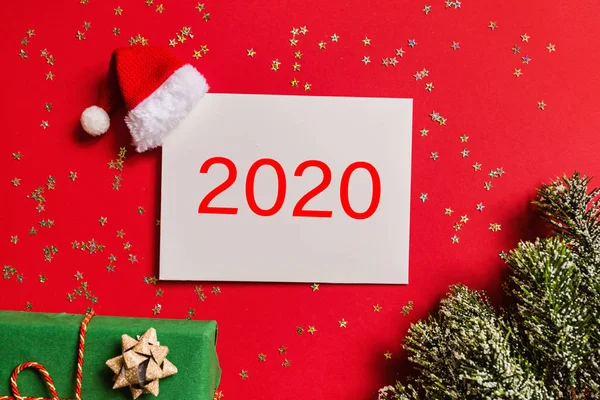 Kosong kertas dengan topi Santa, hadiah, cabang pohon di latar belakang merah dengan emas confetti. Konsep tahun baru. Kartu ucapan, perayaan Natal 2020. Letak datar, templat, tilikan atas, ruang fotokopi, mockup — Stok Foto