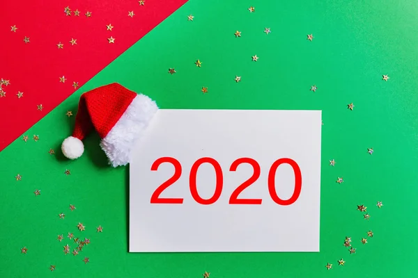 Kosong kertas dengan topi Santa di latar belakang merah dan hijau dengan confetti. Natal dan konsep tahun baru. Kartu ucapan, perayaan Natal 2020. Letak datar, templat, tilikan atas, ruang fotokopi, mockup — Stok Foto