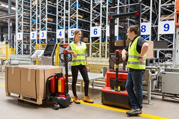 Storehouse employees in uniform standing near pallet truck and forklift in modern automatic warehouse.Boxes are on the shelves of the warehouse. Warehousing, machinery concept. Logistics in stock.