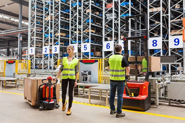 Storehouse employees in uniform standing near pallet truck and forklift in modern automatic warehouse.Boxes are on the shelves of the warehouse. Warehousing, machinery concept. Logistics in stock.