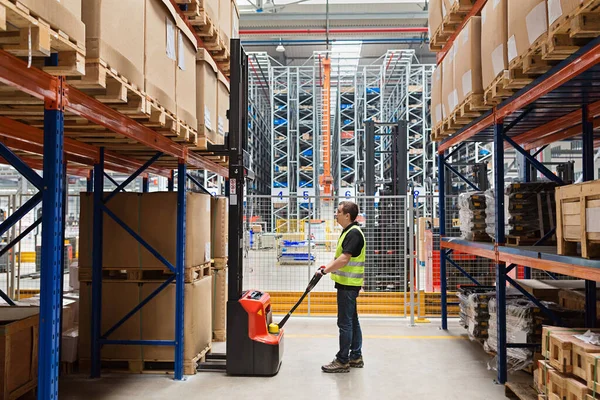 Storehouse medewerker in uniform werkt aan vorkheftruck in modern automatisch magazijn. De dozen liggen op de planken van het magazijn. Opslag, machineconcept. Logistiek in stock. — Stockfoto