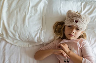 Pijamalı, göz bandı takmış, beyaz yatakta yatan ve anaokuluna gitmeden önce sabahın köründe uyanan güzel sarışın kız manzarası. Uyku zamanı, uyanma ve rahatlama konsepti