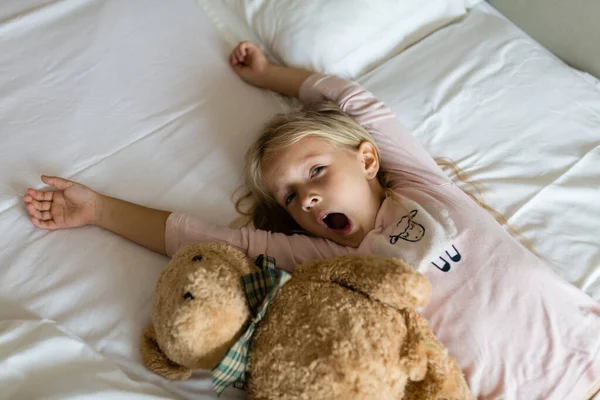 Vista dall'alto della graziosa bambina bionda in pigiama e bendata sdraiata sul letto bianco con orsacchiotto, sveglia la mattina presto prima di andare all'asilo. Tempo di andare a letto, risveglio e relax concetto — Foto Stock