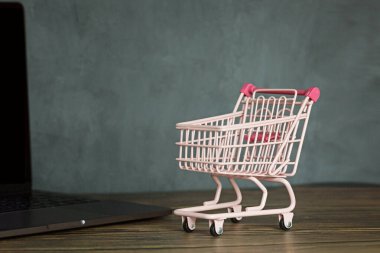 Ahşap masada pembe market arabası ve metin için fotokopi alanı olan dizüstü bilgisayar. Noel için online promosyon alışverişi, Sevgililer Günü, Kara Cuma, siber pazartesi, satış konsepti. Şablon, model