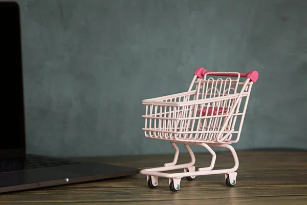 Colore rosa carrello della spesa su scrivania in legno e laptop con spazio copia per il testo. Shopping promozione online per Natale nuovo anno, San Valentino, venerdì nero, cyber lunedì, concetto di vendita. Modello, mockup — Foto Stock