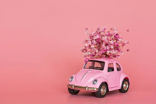 Pink retro toy car delivering bouquet of flowers on pink background. Valentine day, February 14 card. International women day 8 March, mother day, birthday gift. Copy space, mockup, template, minimal