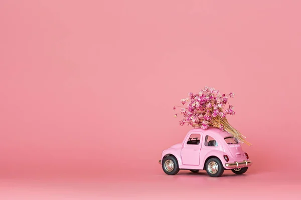 Pink retro toy car delivering bouquet of flowers on pink background. Valentine day, February 14 card. International women day 8 March, mother day, birthday gift. Copy space, mockup, template, minimal