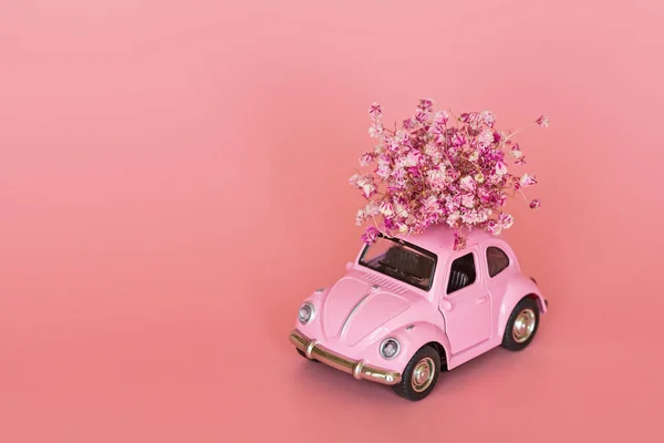 Pink retro toy car delivering bouquet of flowers on pink background. Valentine day, February 14 card. International women day 8 March, mother day, birthday gift. Copy space, mockup, template, minimal