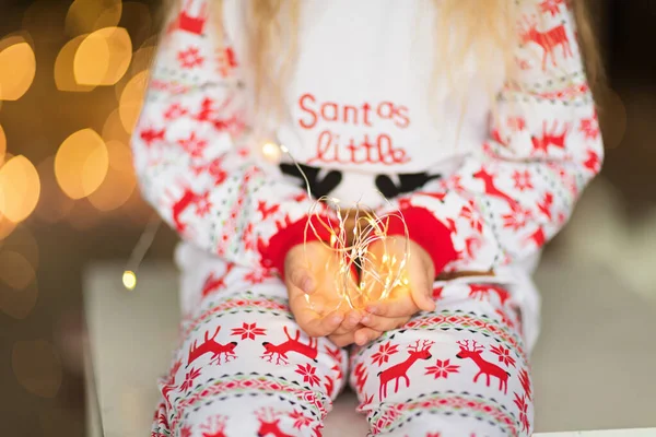 Jolie fille blonde de 5 ans, assise près du sapin de Noël et tenant une guirlande. Chambre décorée avec des guirlandes de Noël. intérieur de la maison, Noël fête, heureux nouveau concept année 2020 — Photo