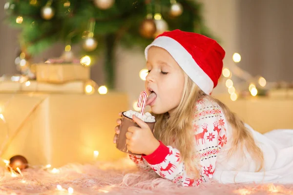 Jolie fille blonde de 5 ans buvant du cacao chaud près du sapin de Noël. Chambre décorée avec des guirlandes de Noël. intérieur de la maison, Noël fête, heureux nouveau concept année 2020 — Photo