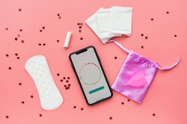 Poşetli regl kupası, hijyenik pedler, tampon, konfetili pembe arka planda adet takvimi olan cep telefonu. O dönemde alternatif kadın hijyen ürünü. Kadın sağlığı kavramı. 